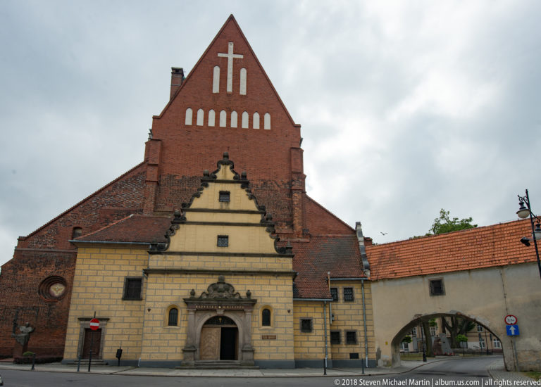 Brick church with steep gable, large cross motif, pale stepped entrance façade, and attached arched passage.