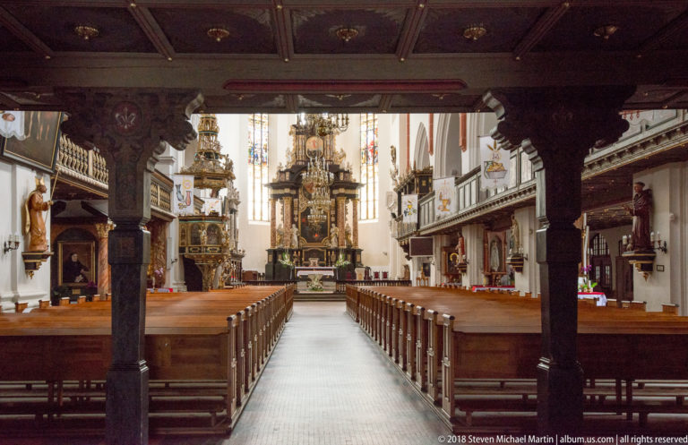 Central aisle of an ornate Oleśnica church with wooden pews, galleries, pulpit, chandeliers, monuments, and a richly decorated altar.