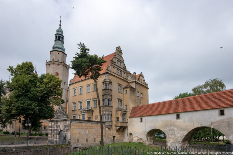 Oleśnica Castle exterior with pale gabled wing, tall tower, corner turret, and a low red-roofed structure with broad arches.