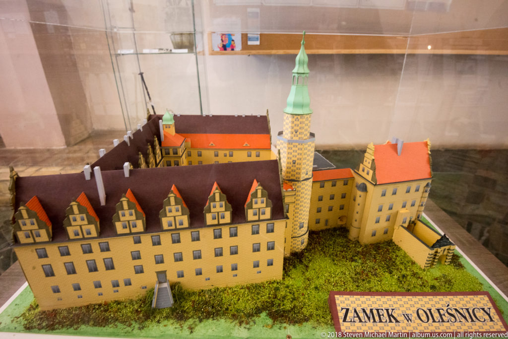 Scale model labeled Zamek Oleśnica with gabled castle wings, red roofs, round tower, and enclosed courtyard.