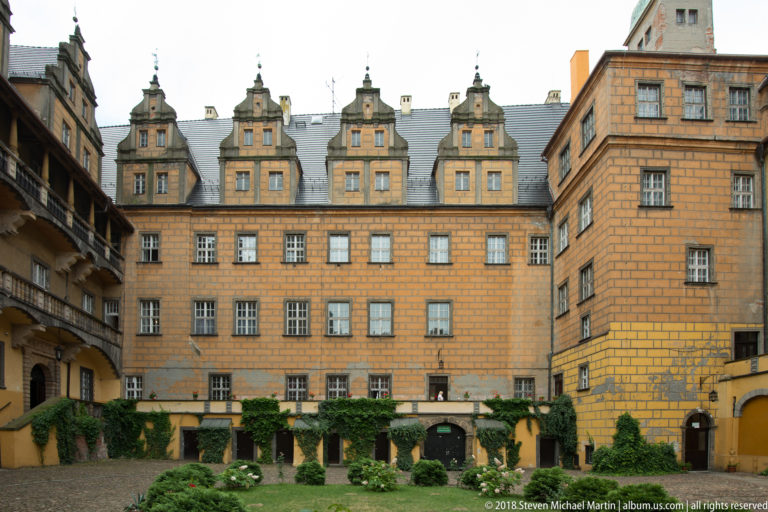 Symmetrical patterned castle façade with four ornate gables, rows of windows, ivy, and arched ground-level openings.