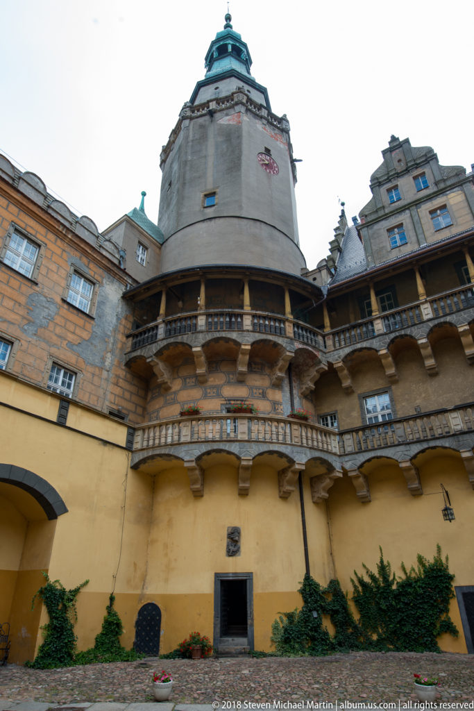 Round pale tower with green roof and projecting gallery rising above the yellow walls of Oleśnica Castle courtyard.