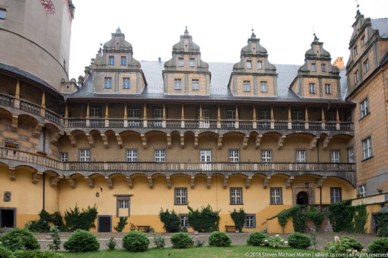 Frontal view of a castle courtyard wing with two gallery levels, projecting supports, ornate gables, windows, and clipped shrubs.