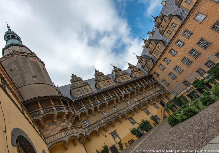 Angled upward view of Oleśnica Castle courtyard with arcaded galleries, ornate gables, patterned façades, and a round tower.