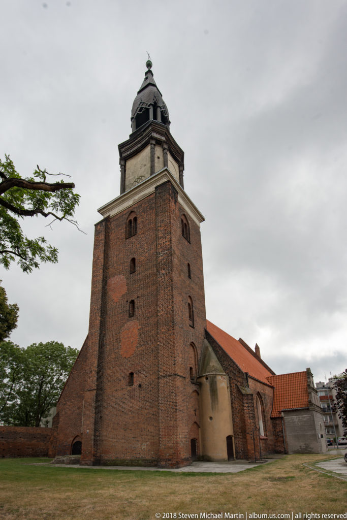 Massive brick medieval church tower with dark octagonal top and cupola beside red-roofed brick naves in Oleśnica.