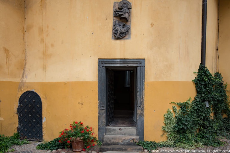 Dark stone-framed doorway in a yellow wall beside ivy, red flowers, and a small studded arched door.