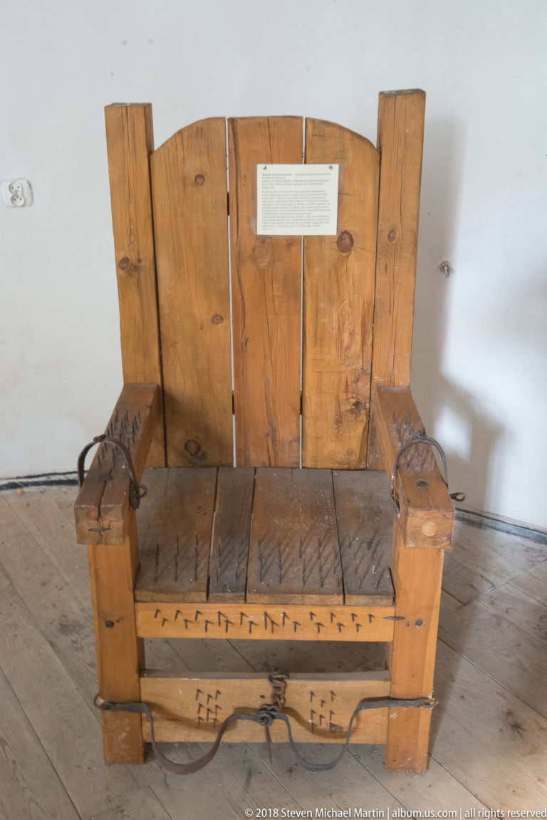 Heavy wooden restraint chair with leather straps, metal fastenings, broad plank back, and marked lower rail.