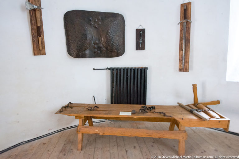 Wooden bench holding assorted iron and wooden implements beneath wall-mounted historic objects in a plain castle room.