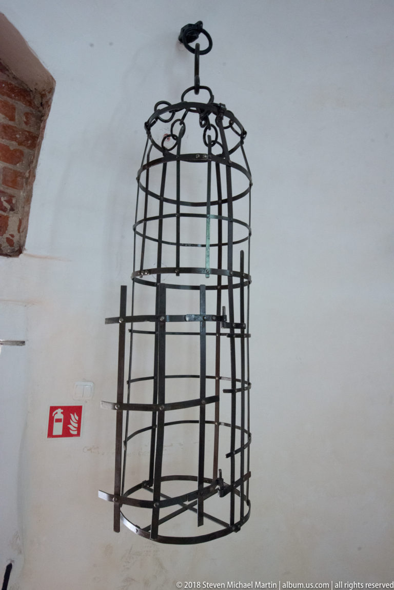Suspended black iron cage with curved vertical bars and circular bands displayed against a white castle wall.
