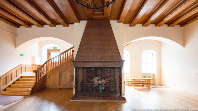 Large hooded brick fireplace beside a wooden staircase in a bright room with arched windows and beamed ceiling.