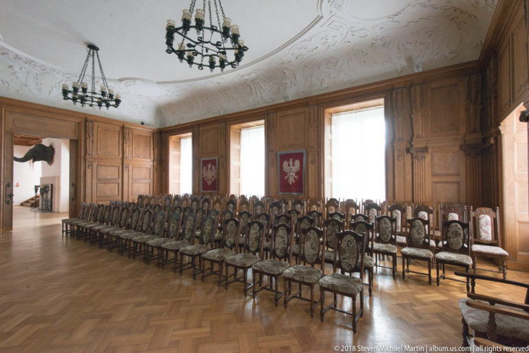 Wood-paneled castle hall with rows of carved chairs, parquet floor, chandeliers, tall windows, and heraldic banners.