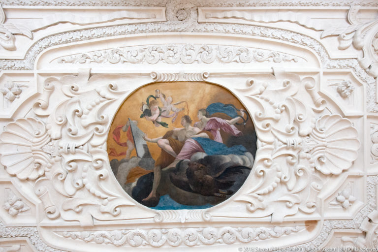 Oval figurative ceiling painting with figures among clouds, surrounded by elaborate white stucco scrollwork and molded ornament.