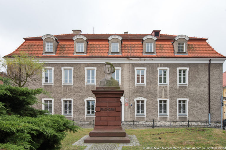 Bust of Fryderyk Chopin on a tall reddish pedestal before the gray former Widows’ House and music school in Oleśnica.