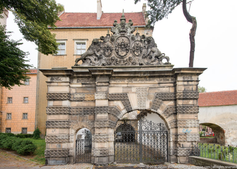 Rusticated stone gateway with wrought-iron gates, carved crest, scrolling ornament, and a smaller side entrance.