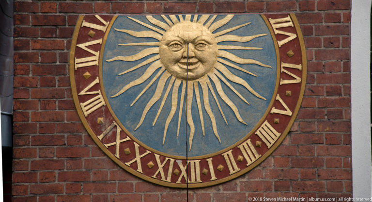 Round decorative sundial with a golden sun face, radiating rays, blue field, and Roman numerals on brick.