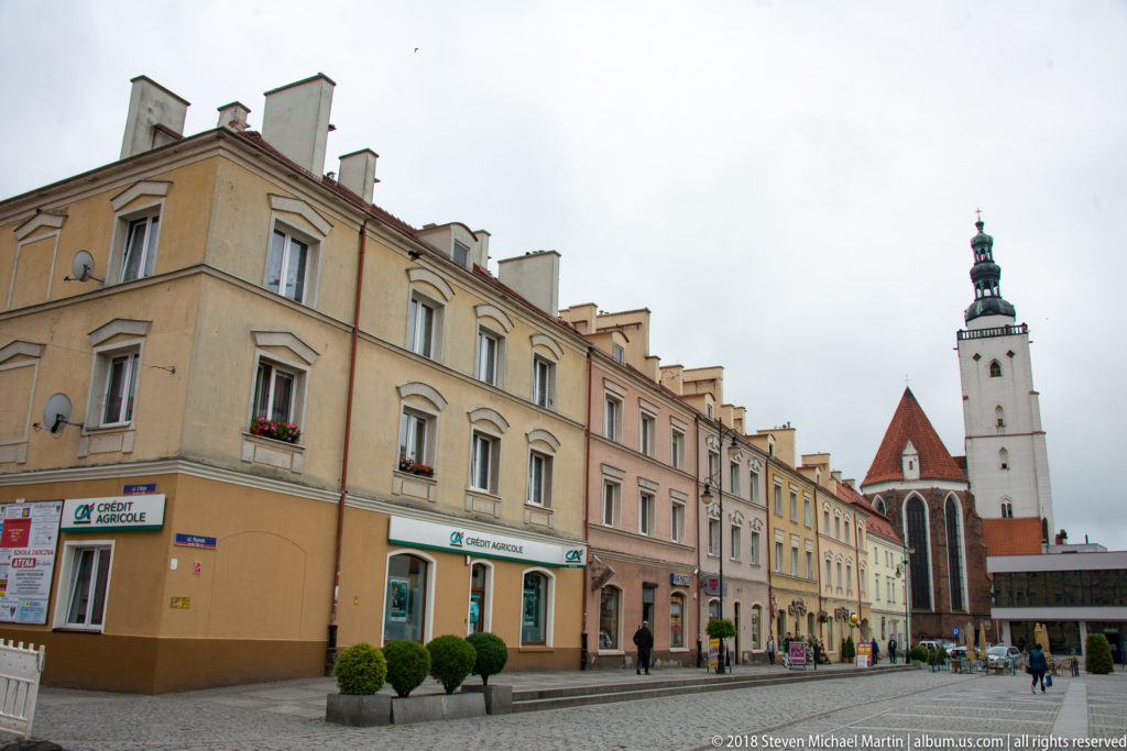Pastel townhouses and shops line an Oleśnica street leading toward a tall church tower and red-roofed church.