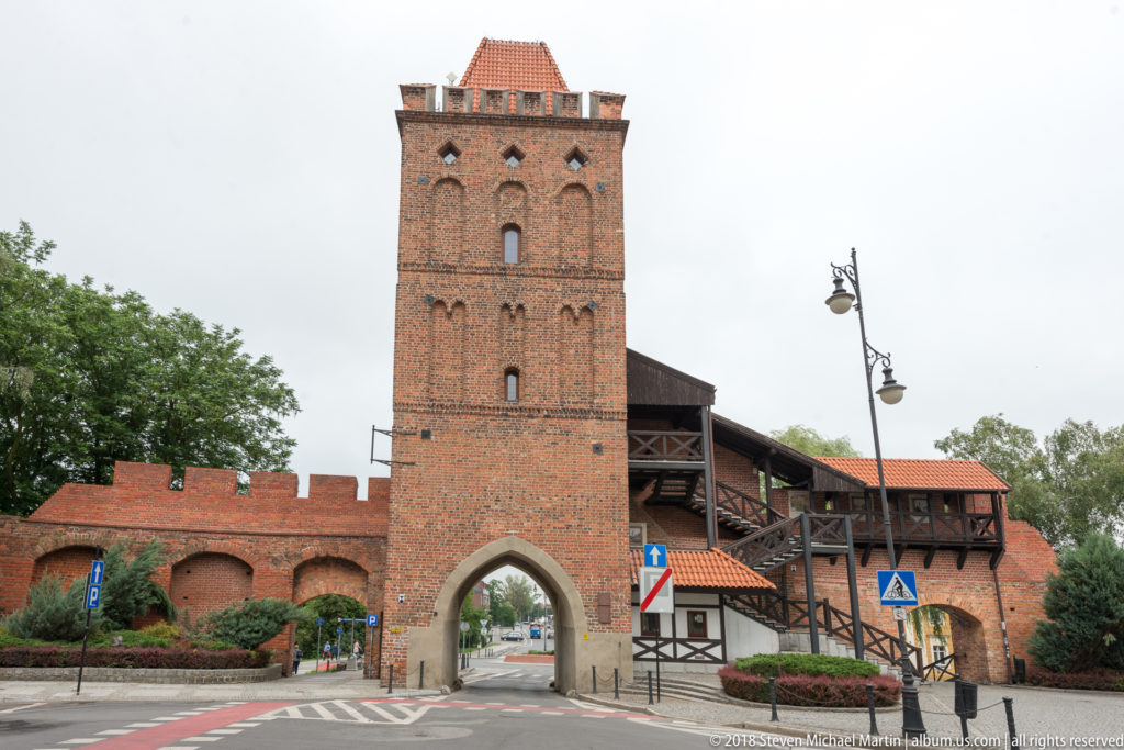 Tall brick gate tower with pointed road passage, crenellated walls, and timber gallery beside a modern street.