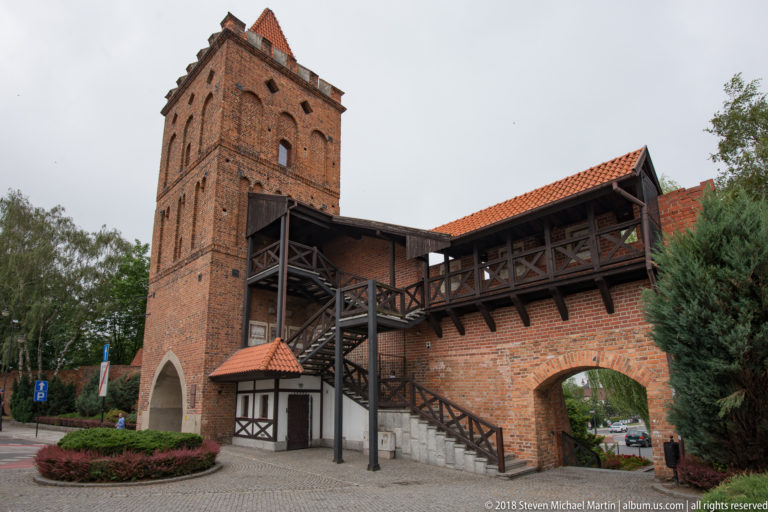 Brick gate tower beside a covered timber wall-walk and exterior wooden stairs in Oleśnica.