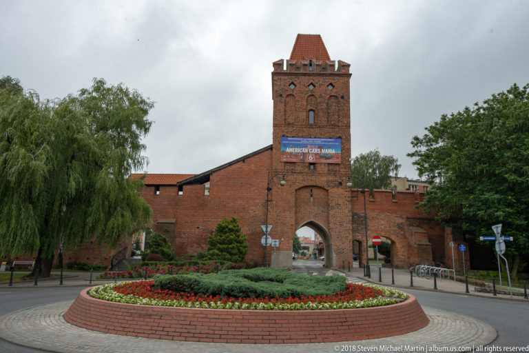 Brick medieval gate tower with pointed passage, crenellated walls, landscaped roundabout, and trees in Oleśnica.