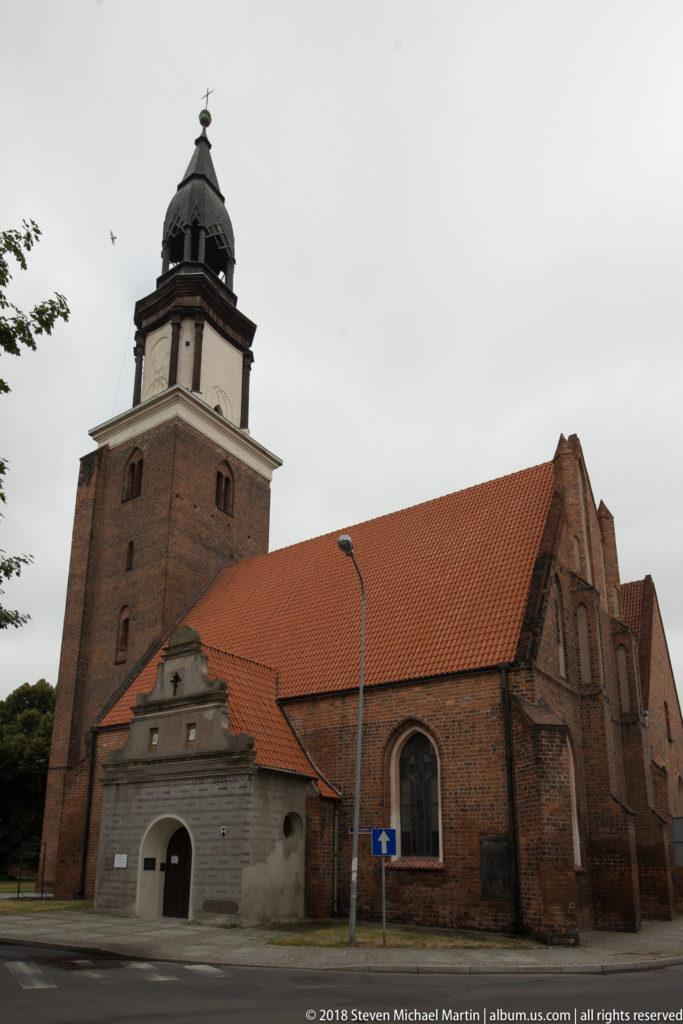 Brick Orthodox church complex in Oleśnica with massive square medieval tower, dark cupola, red tiled roofs, and Gothic windows.