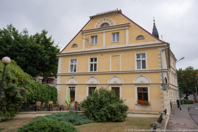 Yellow former Widows’ House in Oleśnica, now a music school, with symmetrical windows, high roof, shrubs, and garden seating.