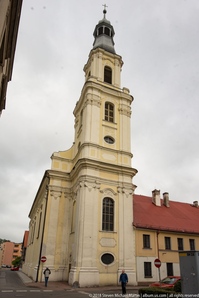 Pale Baroque Holy Trinity Church in Oleśnica with a tall tiered tower, dark cupola, pilasters, and red-roofed nave.
