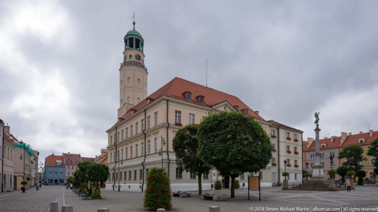 Oleśnica town hall with pale façade, red roof, tall clock tower and green cupola beside clipped trees in the market square.