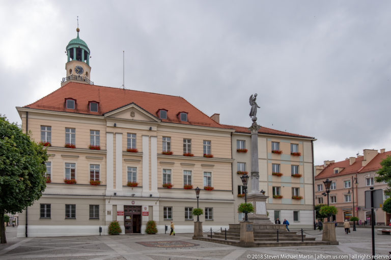 Stone column monument topped by a figure in Oleśnica’s market square, with pale historic buildings and clipped trees behind.