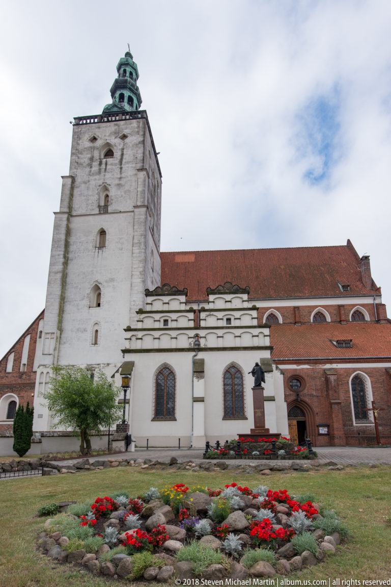 Large brick church with massive pale tower, green cupola, red tiled roof, white side addition, and flower beds.