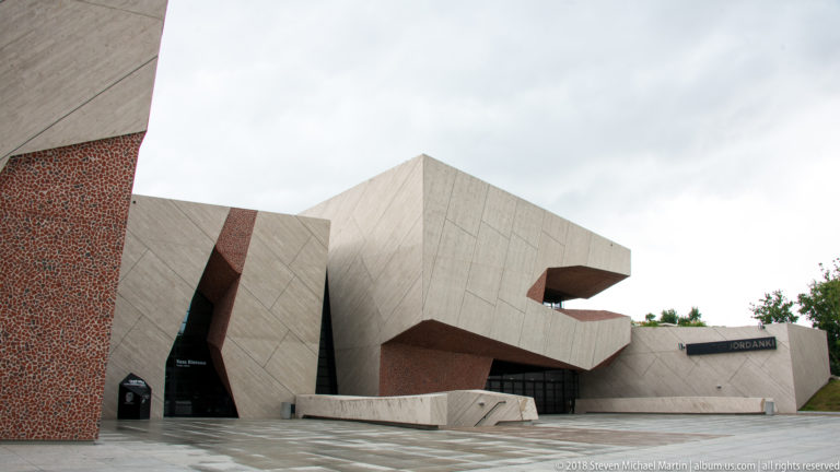 Angular pale-concrete cultural center with reddish aggregate walls, deep recessed openings, and wet paving under an overcast sky.