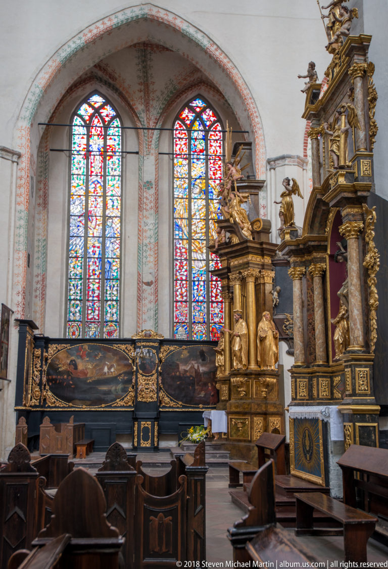Two tall stained-glass windows beside an elaborate gilded Baroque altar, with dark wooden seating in the foreground.