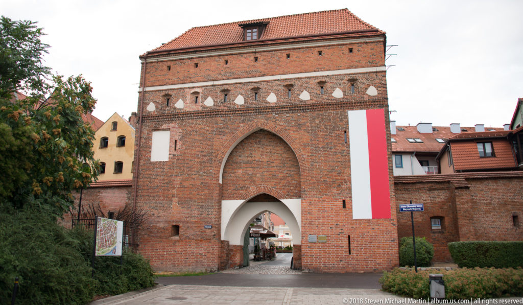 Massive brick medieval gate with pointed arch, white decorative motifs and Polish flag in Toruń.