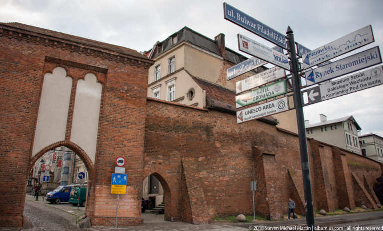 Brick medieval wall and arched gateway beside Old Town directional signs in Toruń.