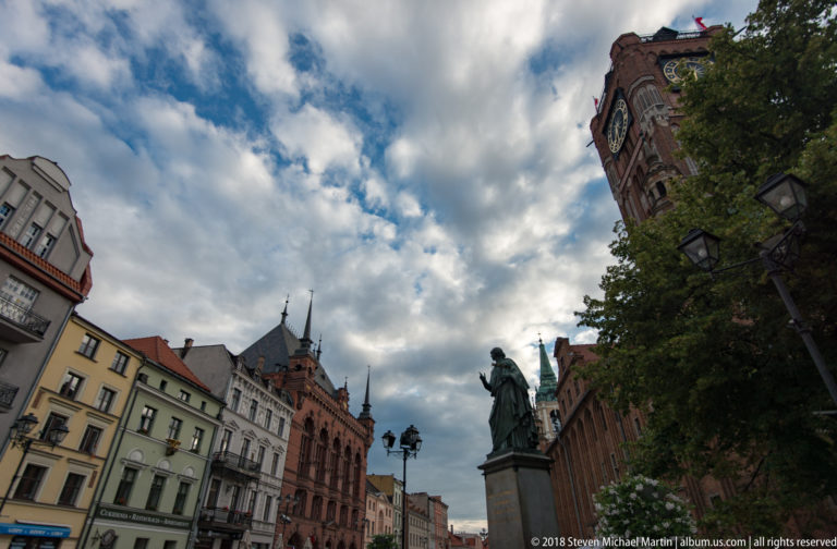 Low-angle view across Toruń market square toward Old Town Hall tower, monument and historic façades.