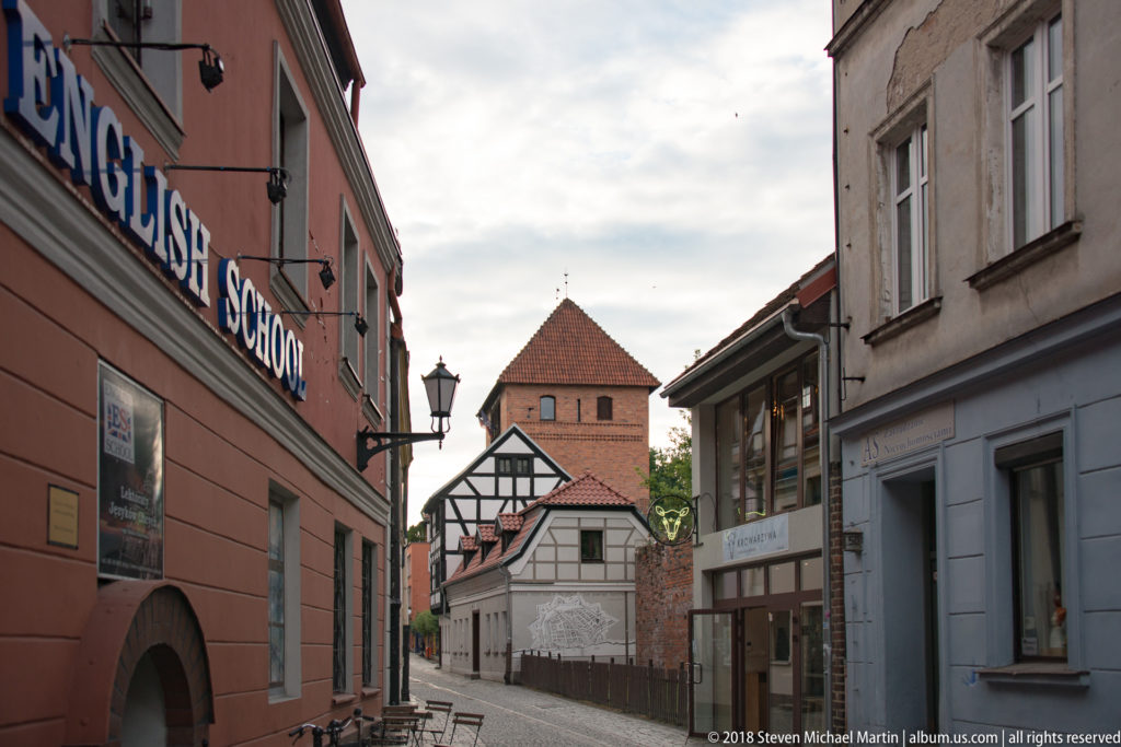 Narrow Toruń street lined with historic buildings and leading toward a square brick defensive tower.