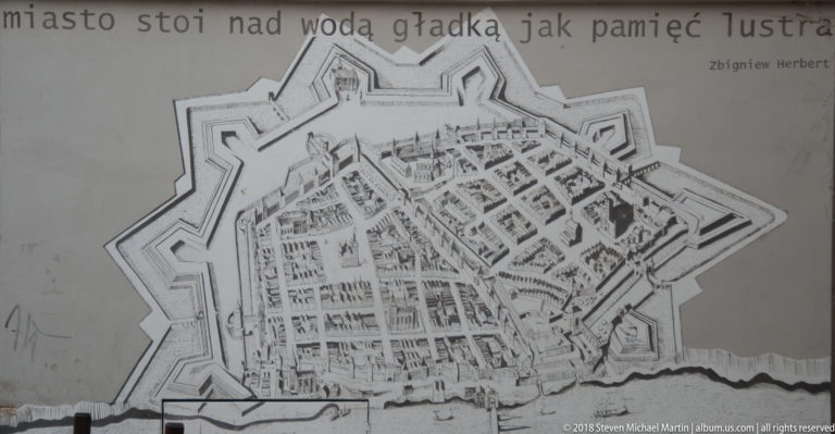 Monochrome wall mural showing an overhead map of historic Toruń enclosed by fortifications.