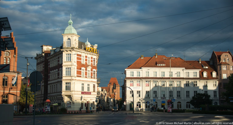 Historic corner hotel with turret and rooftop sign on a broad Toruń street under overhead wires.