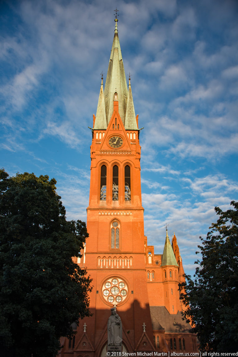 Red-brick Church of St. Catherine in Toruń with tall Neo-Gothic tower, rose window and copper spire.