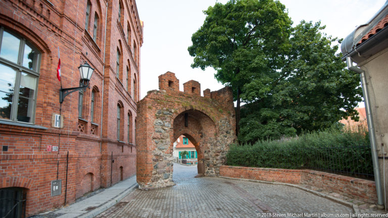 Small arched gateway in weathered brick-and-stone wall opening onto a cobbled Toruń lane.