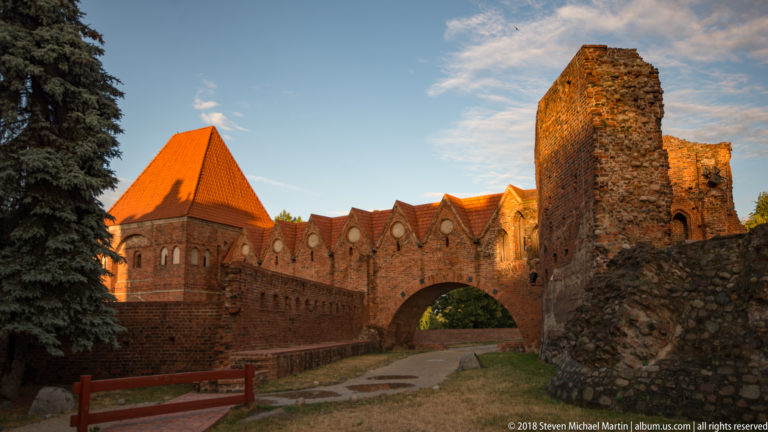 Sunlit brick ruins of Toruń’s Teutonic Castle with pointed arch, gabled wall and tall ruined tower.