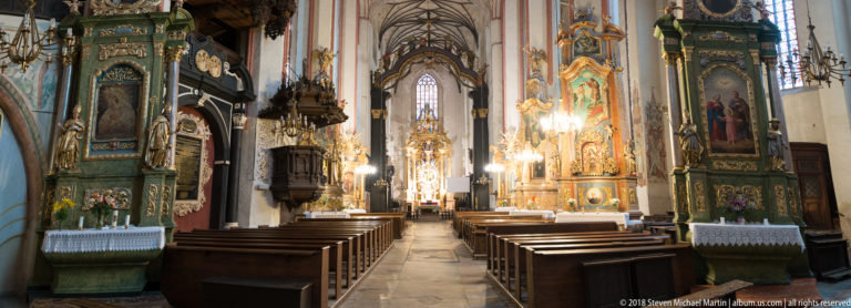 Gothic church nave lined with ornate gilded side altars and leading toward a bright high altar.