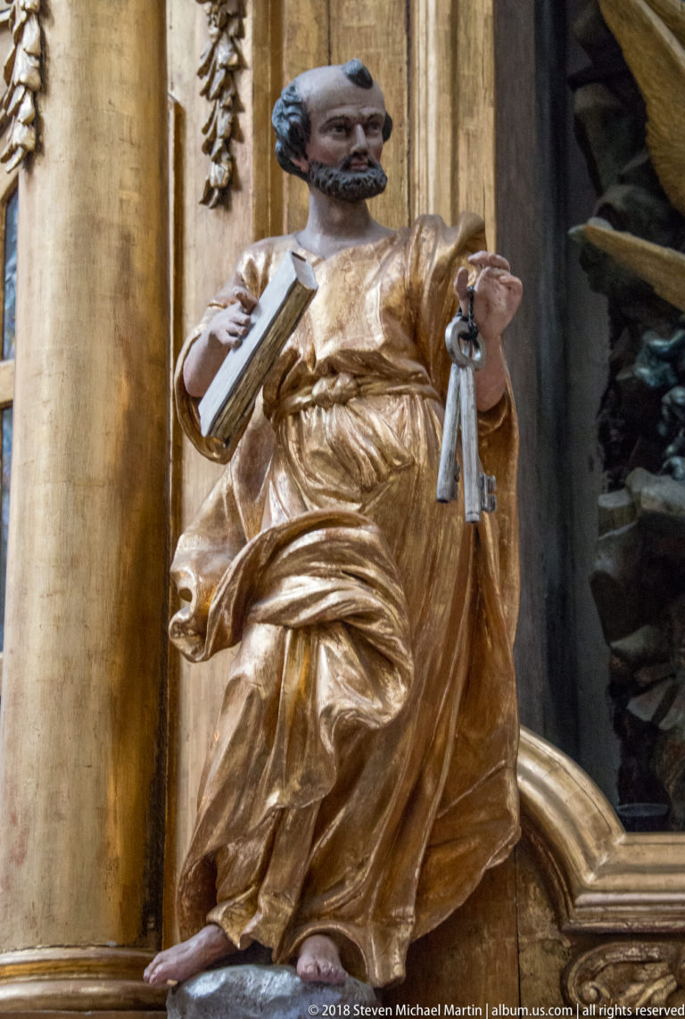 Carved bearded saint in a gilded robe holding large keys beside a Baroque altar column.