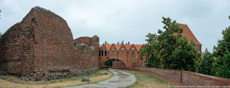 Path between tall ruined brick walls leading toward a gabled arch at Toruń’s Teutonic Castle.