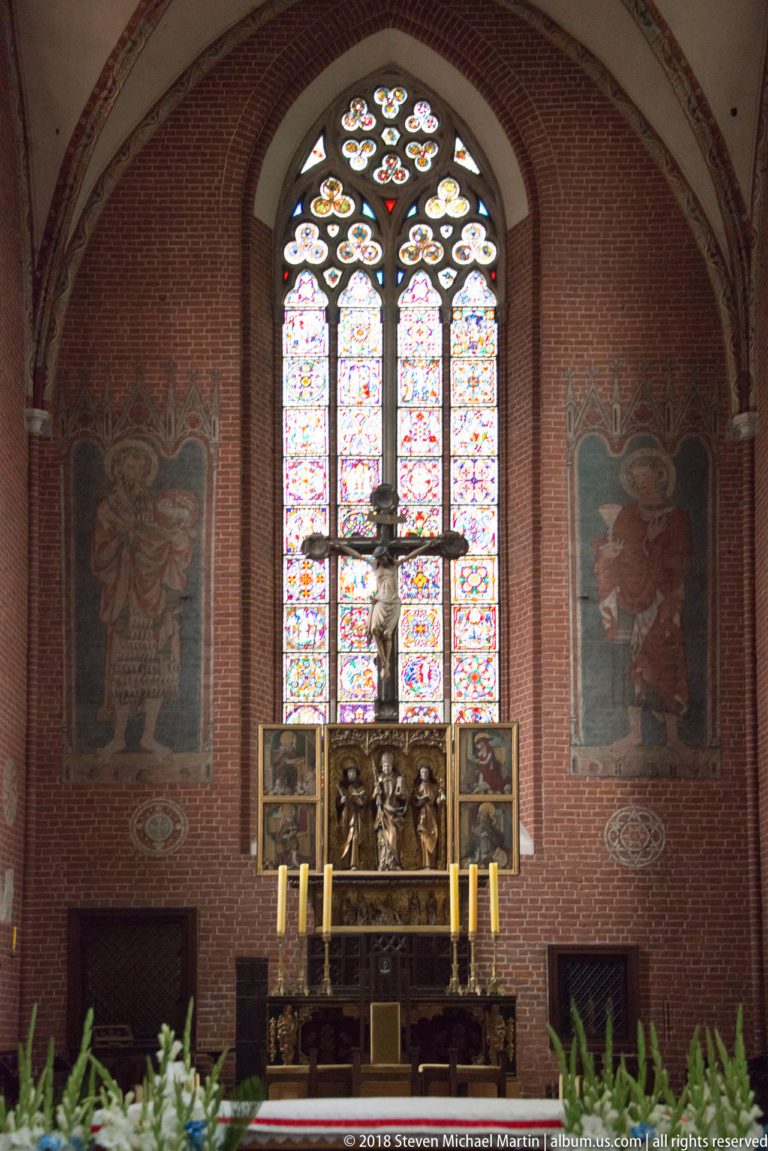 High altar centered beneath a tall stained-glass window on a red-brick wall with large painted figures to either side.