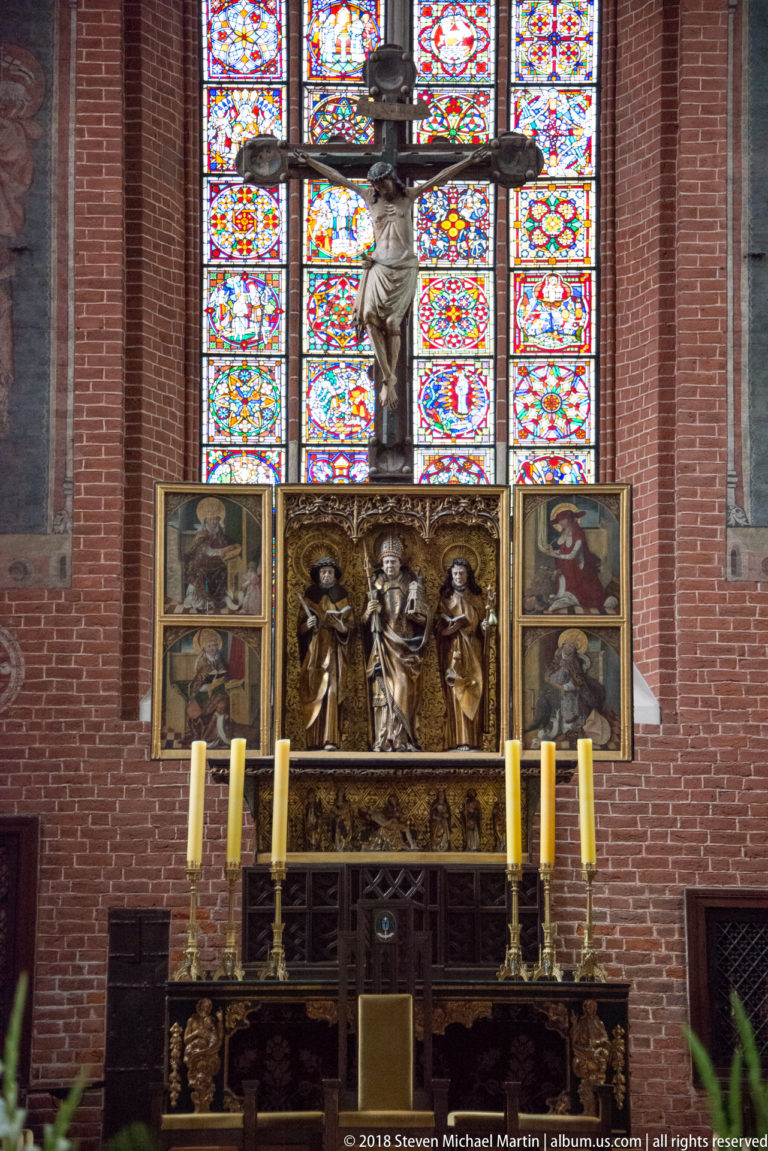 Crucifix above a carved and painted high altar, backed by a large stained-glass window and flanked by tall candles.