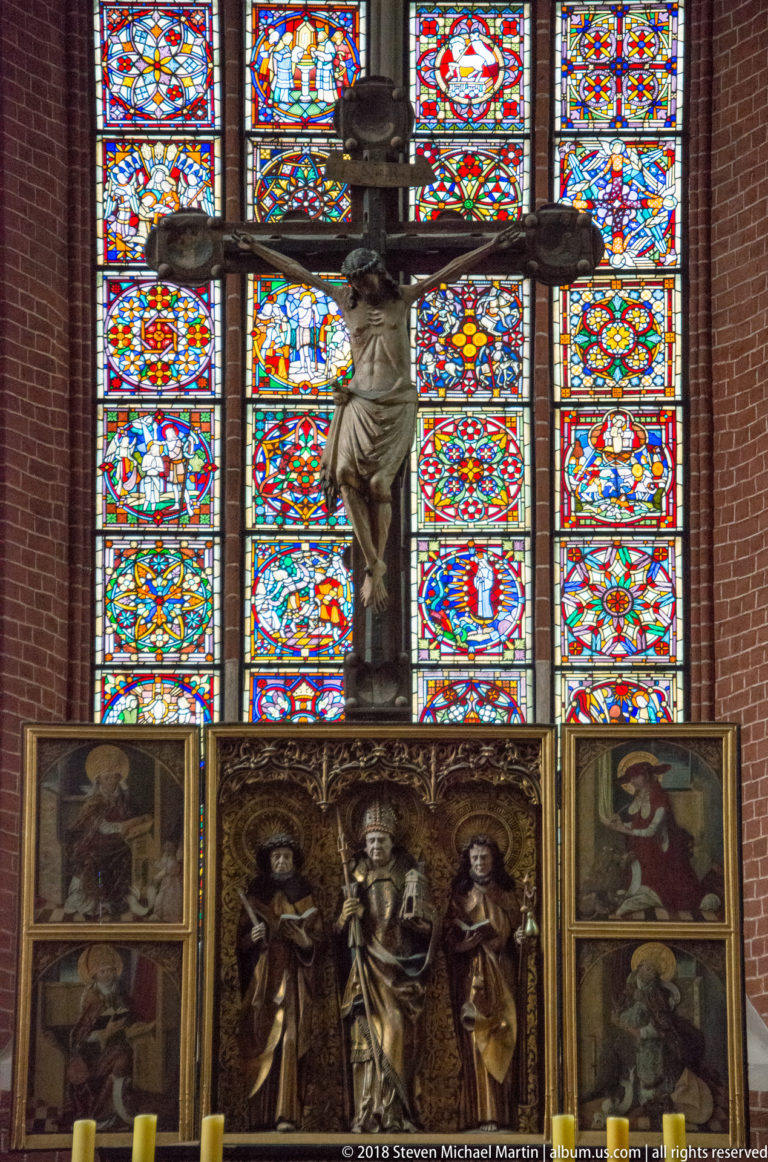 Large crucifix silhouetted against colorful stained glass above carved and painted figures of the high altar.