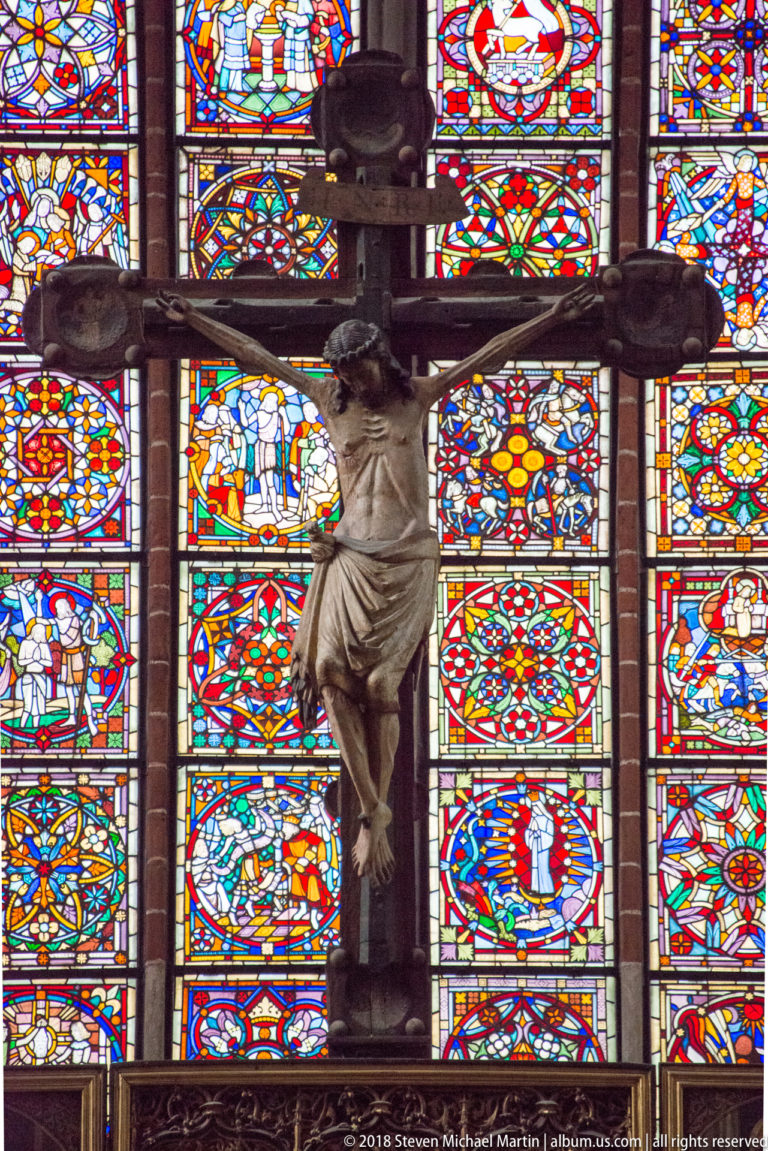 Close view of a sculpted Christ on a dark wooden cross set against brightly colored stained-glass panels.