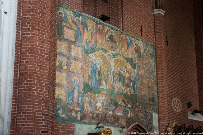 Large faded multi-scene mural painted directly on a red-brick church wall, with figures arranged in several compartments.
