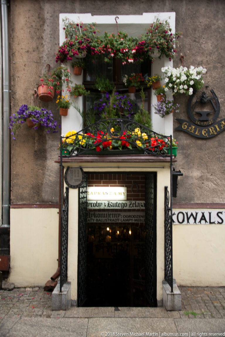 Narrow shop entrance beneath a historic façade, surrounded by dense window boxes and planters filled with colorful flowers.