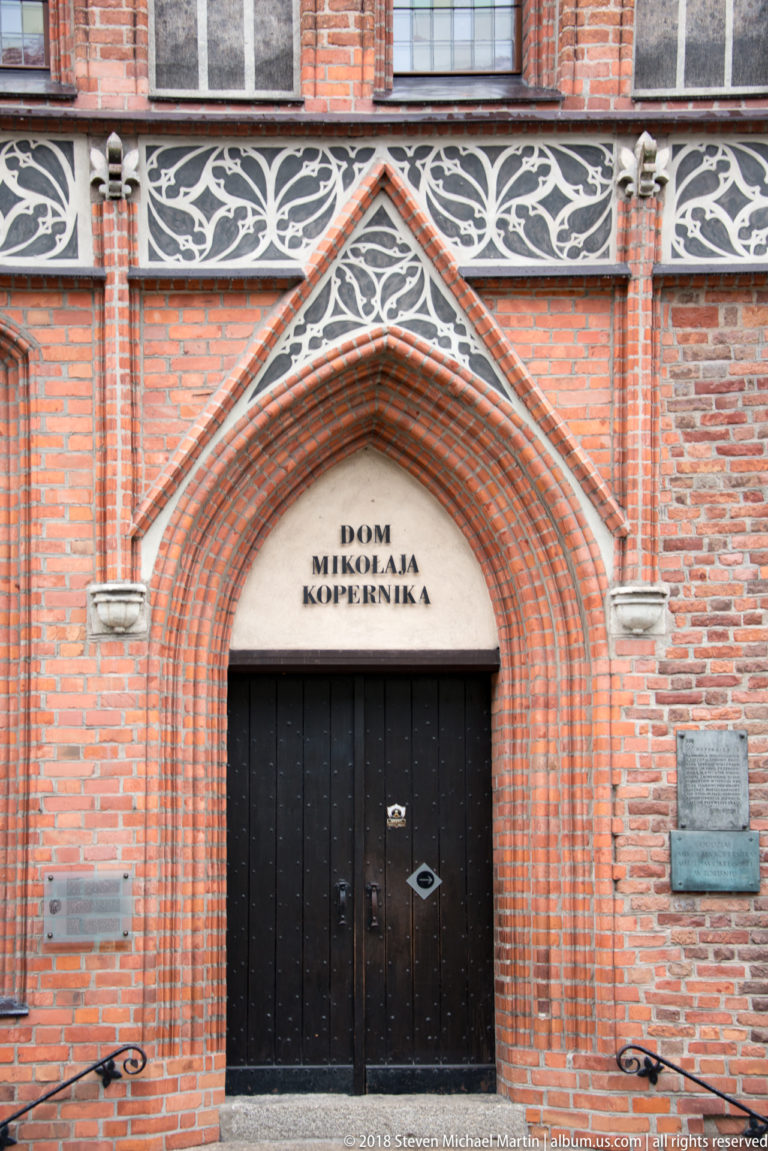 Pointed Gothic brick doorway with black double doors and the inscription “Dom Mikołaja Kopernika” above the entrance.